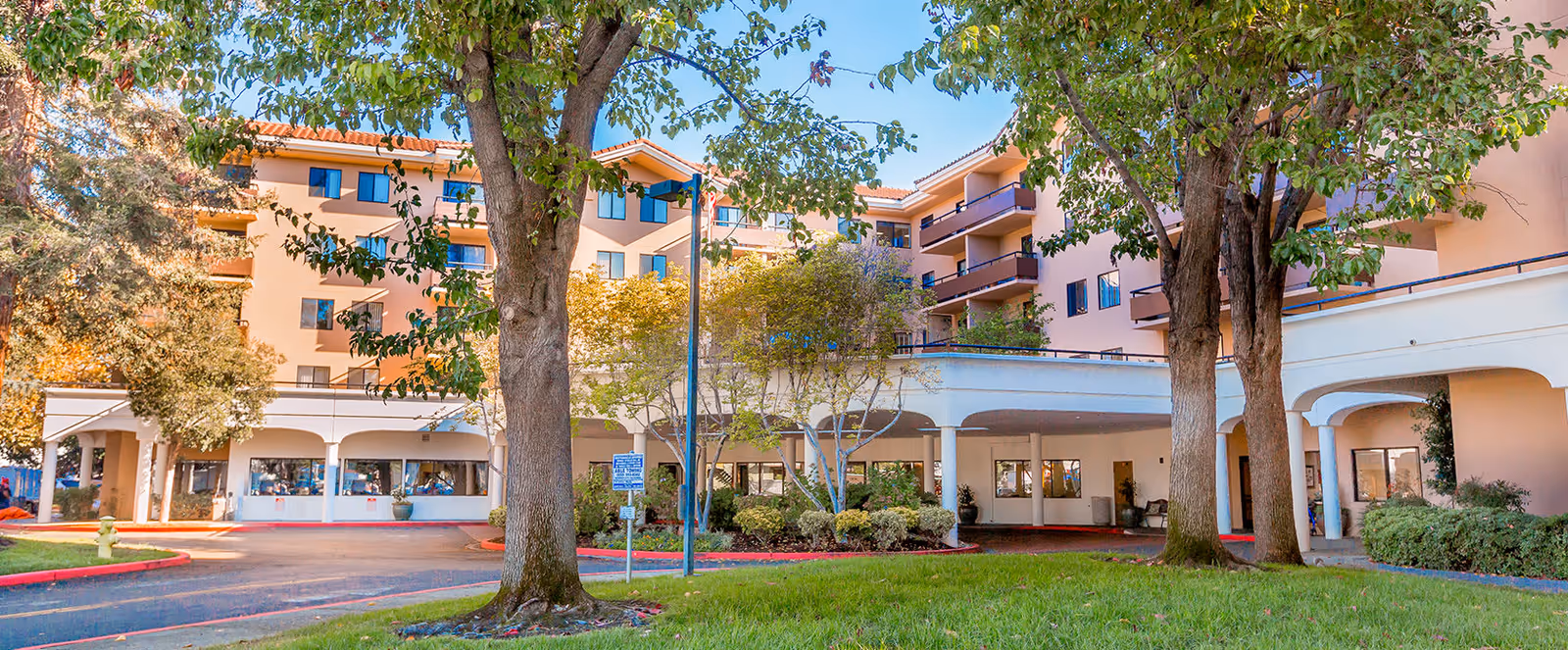 Exterior view of a multi-story senior living facility with peach-colored walls and balconies, surrounded by trees and greenery, with a driveway and covered entrance area.