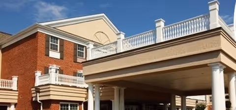 Exterior view of a brick building with white columns and railings, featuring a covered entrance area under a balcony with decorative balusters, under a clear blue sky.