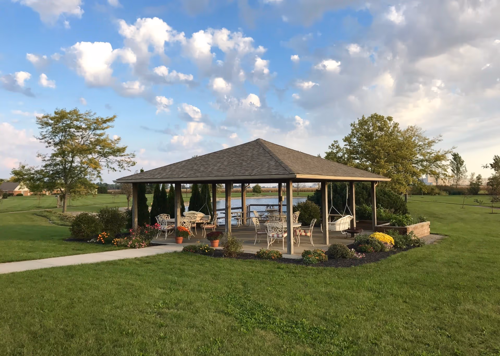A wooden gazebo with a shingled roof situated in a grassy area with trees and a pond in the background. The gazebo contains several metal chairs and tables, potted flowers, and two white swings hanging from the roof. The sky is partly cloudy with patches of blue.