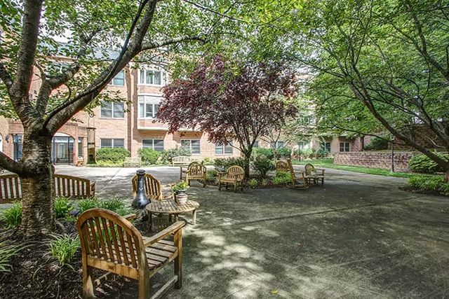 Outdoor courtyard area at a senior living facility with wooden benches and chairs arranged around small tables under large leafy trees, surrounded by brick buildings.