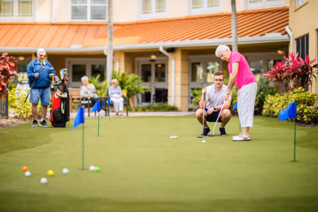 An elderly woman is playing putting golf on a green putting area outside a senior living facility. A younger man is crouched beside her, assisting with her putt. In the background, an elderly man with a golf bag and two seated elderly women are watching. The building of the facility is visible behind them with plants and a covered walkway.