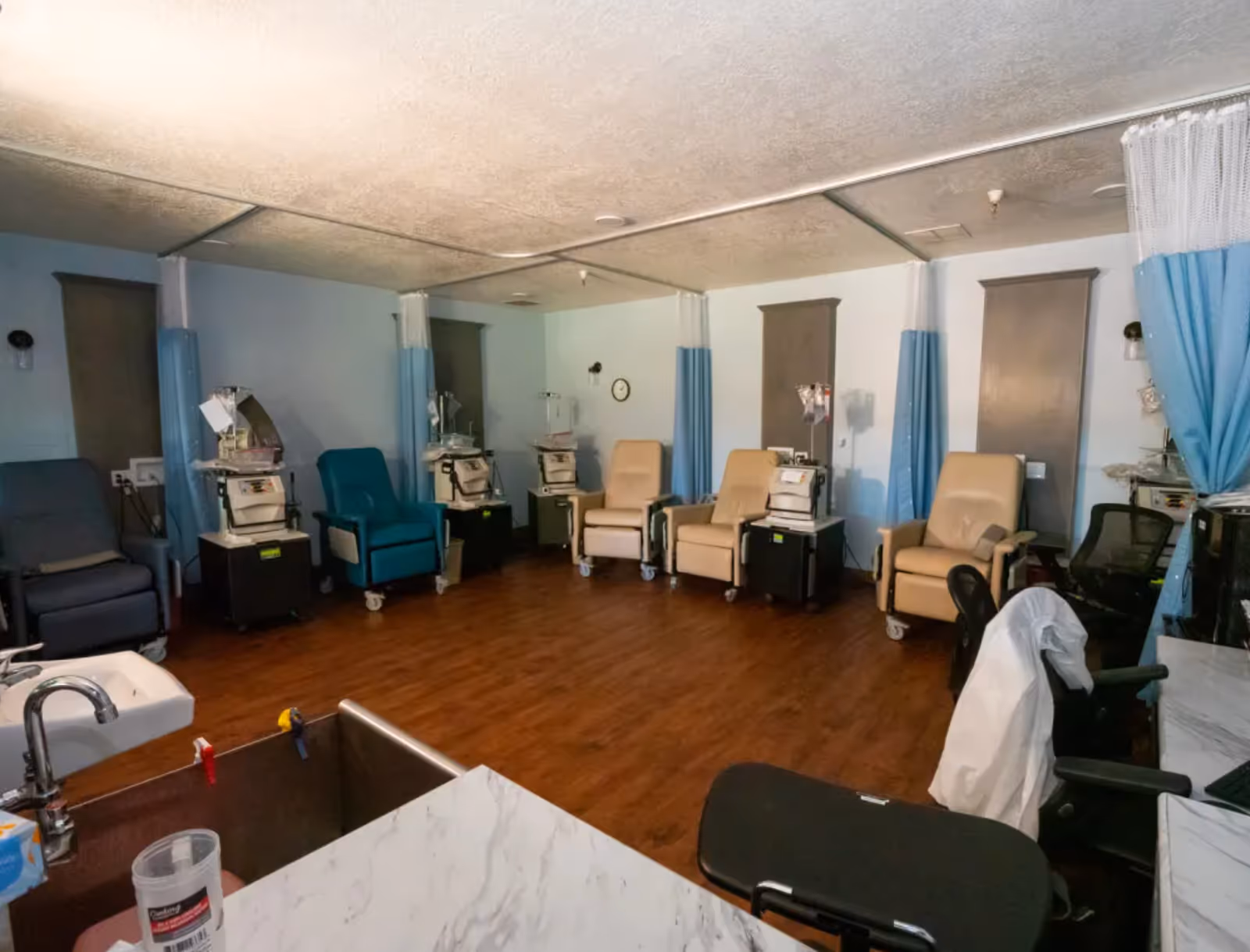 A medical treatment room with several reclining chairs arranged along the walls, each paired with medical equipment such as IV stands and dialysis machines. The room has light blue walls, wood flooring, privacy curtains, and a sink with a countertop in the foreground.