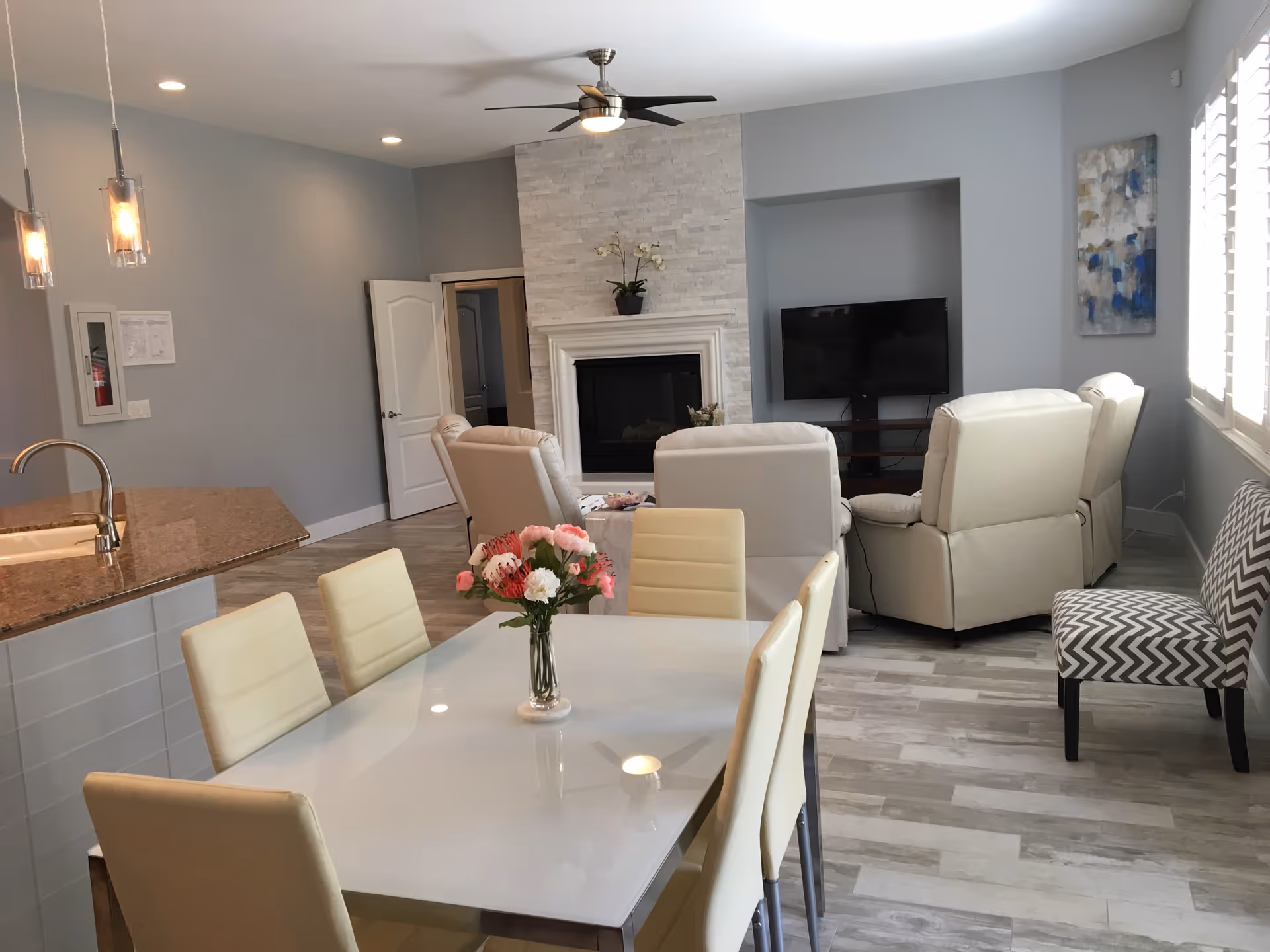A modern senior living common area featuring a dining table with six cream-colored chairs and a vase of flowers in the center. In the background, there is a living room with four beige recliner chairs arranged around a fireplace with a stone surround and a flat-screen TV mounted on the wall. The room has light gray walls, wood-look tile flooring, a ceiling fan, and large windows with white blinds allowing natural light to enter.