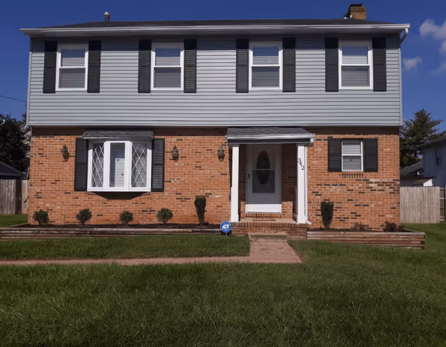 Two-story residential building with a brick lower half and gray siding upper half, featuring multiple windows with black shutters, a white front door with a small porch, and a well-maintained lawn with a walkway leading to the entrance.