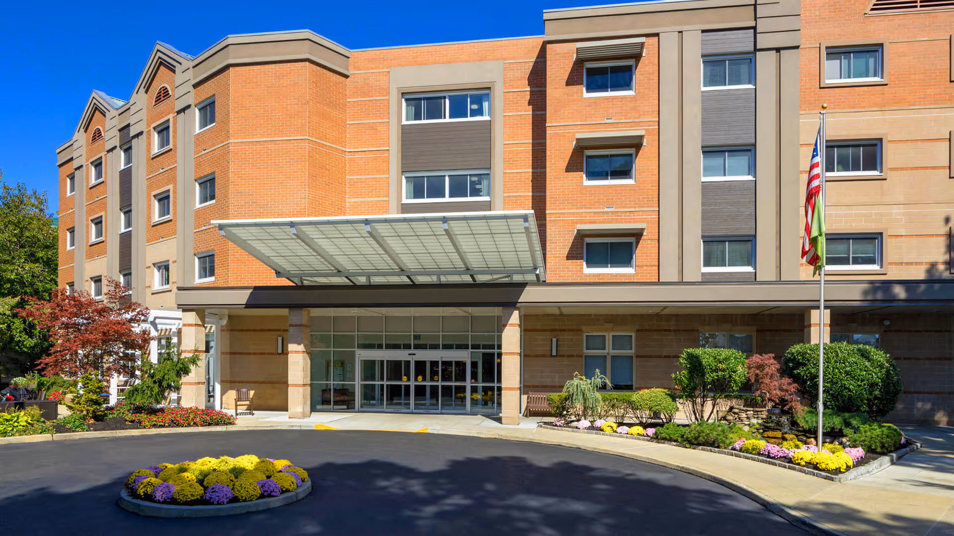 Front exterior view of a multi-story senior living facility building with a covered entrance, landscaped flower beds, shrubs, and an American flag on a flagpole under a clear blue sky.