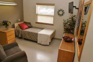 A compact resident bedroom featuring a single bed, dresser, window, and seating area.
