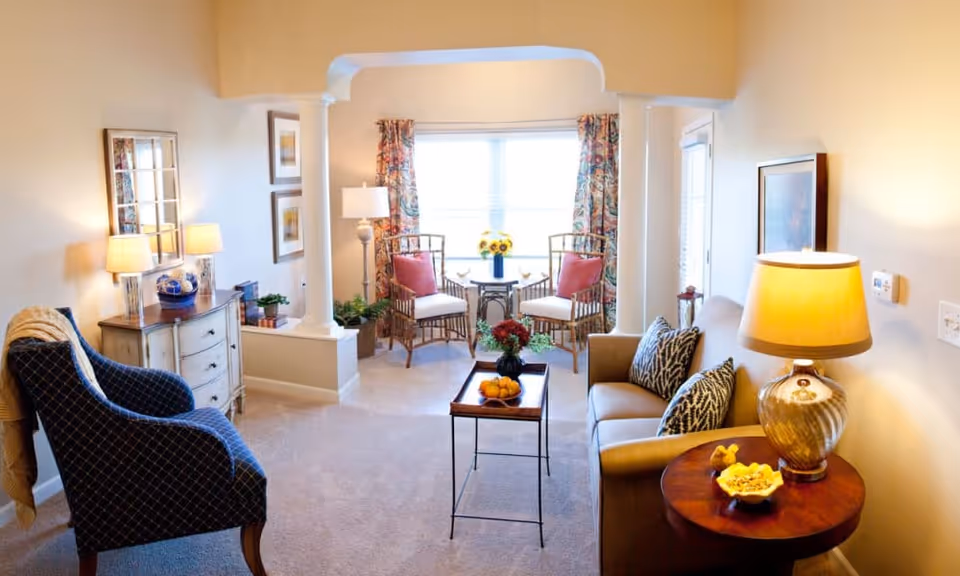 A cozy living room with a beige sofa adorned with patterned pillows, a wooden side table with a lamp and decorative items, a blue upholstered armchair with a blanket draped over it, a small wooden coffee table with a bowl of fruit and flowers, and two wicker chairs with cushions near a window with floral curtains. The room is warmly lit and decorated with framed artwork and plants.