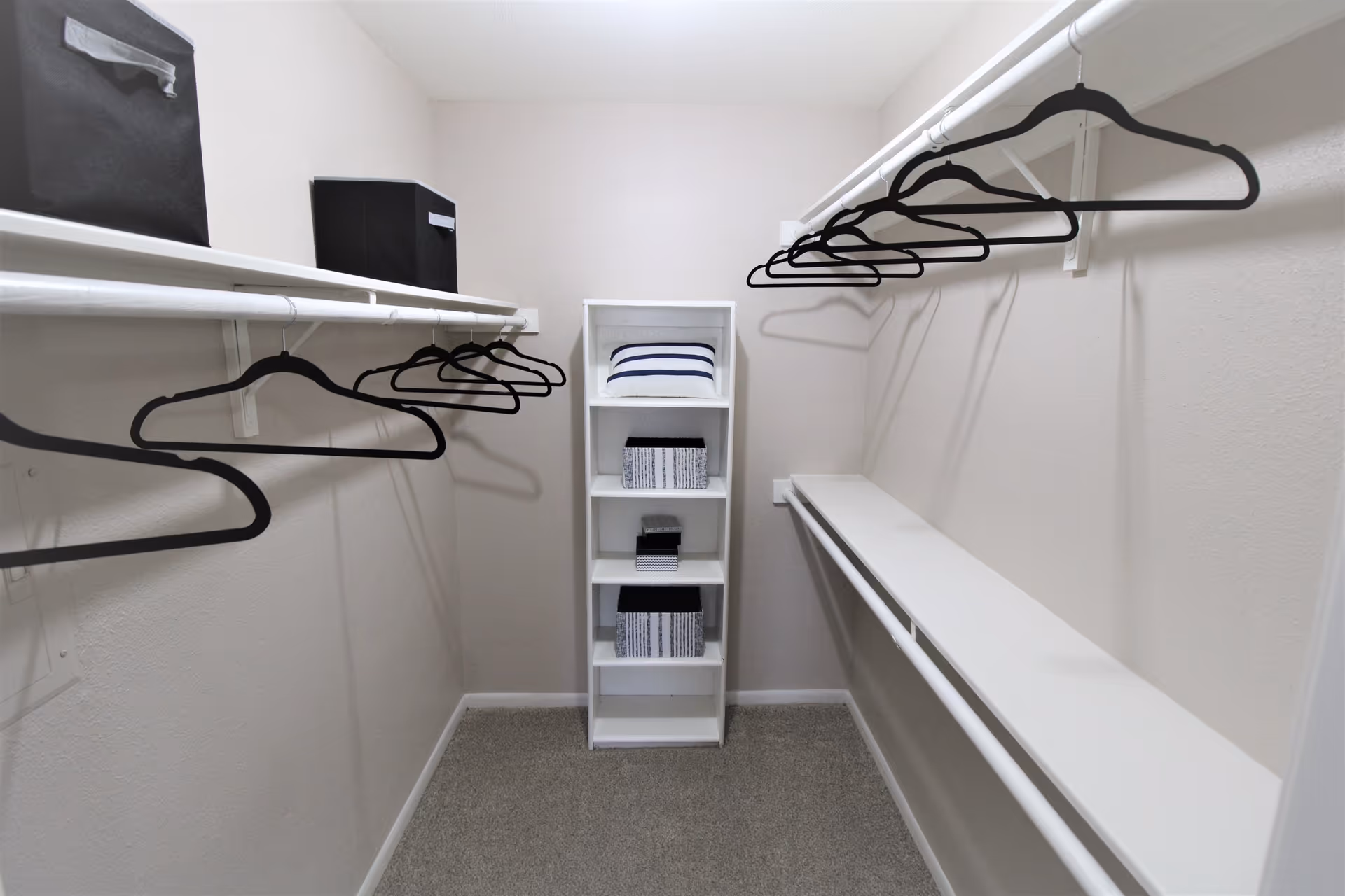 A walk-in closet with white walls and carpeted floor. The closet has two white hanging rods on either side with black hangers. At the end of the closet, there is a white shelving unit holding folded towels and storage boxes.