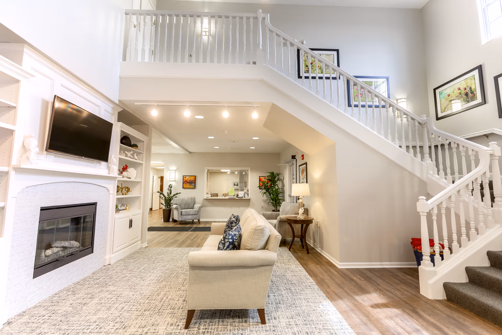 A bright and spacious living room area in a senior living facility featuring a beige sofa with patterned cushions, a fireplace with a mounted flat-screen TV above it, built-in white shelves, a staircase with white railings leading to an upper floor, and framed artwork on the walls. The room has wood flooring and a large area rug, with additional seating and plants visible in the background.