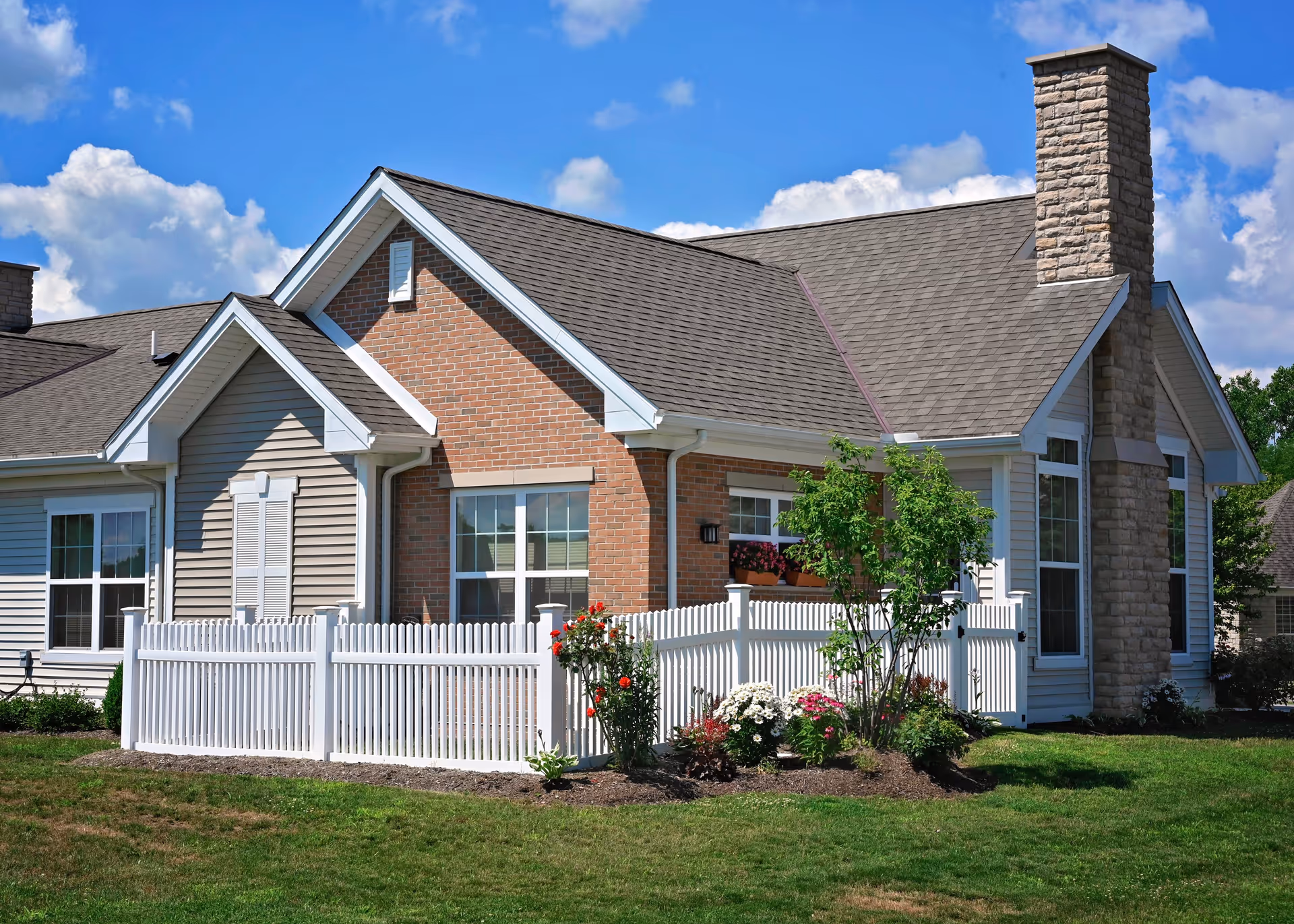 Exterior view of a single-story residential building with a combination of brick and siding walls, a gray shingled roof, a stone chimney, white-framed windows, and a white picket fence surrounding a small garden with flowers and shrubs under a blue sky with some clouds.