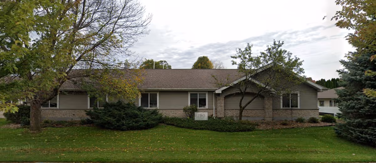 Single-story building with beige siding and a brown roof, surrounded by green grass, bushes, and several trees with some leaves turning yellow, under a cloudy sky.