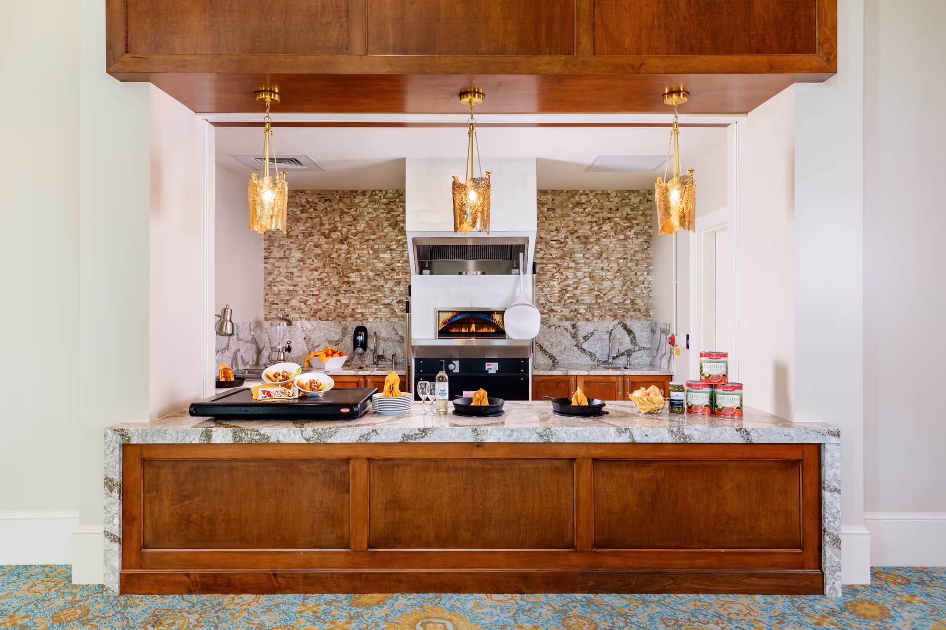 A kitchen area with a marble countertop and wooden cabinetry. Three pendant lights hang above the counter. On the counter, there are plates with food, napkins, and canned goods. The back wall features a stone backsplash and a built-in oven with a pizza peel hanging on the side.