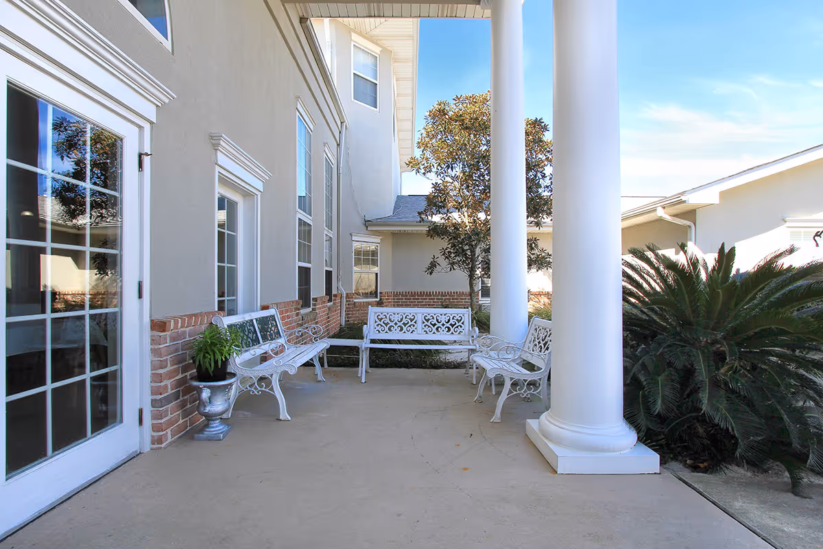 Outdoor covered patio area at Gulf Breeze Courtyard Assisted Living with white decorative metal benches, large white columns, potted plant, and surrounding greenery including a tree and bushes under a clear blue sky.