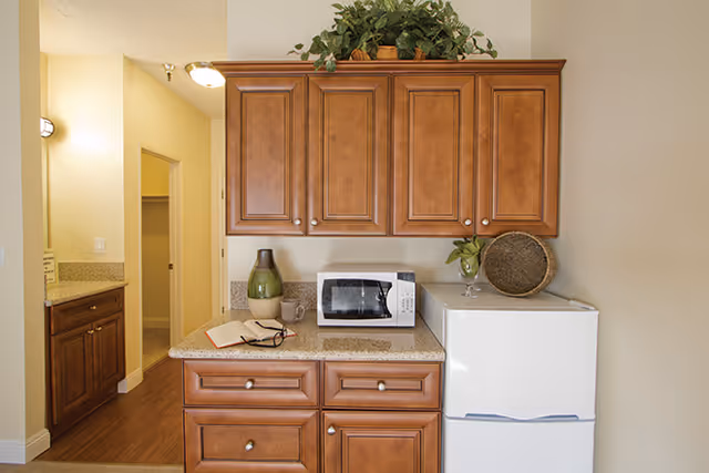 A small kitchen area with wooden cabinets, a white microwave, a white mini refrigerator, and decorative items including a green vase, a book, and a woven basket on the countertop. There is a hallway visible to the left with additional cabinetry and a light fixture on the ceiling.