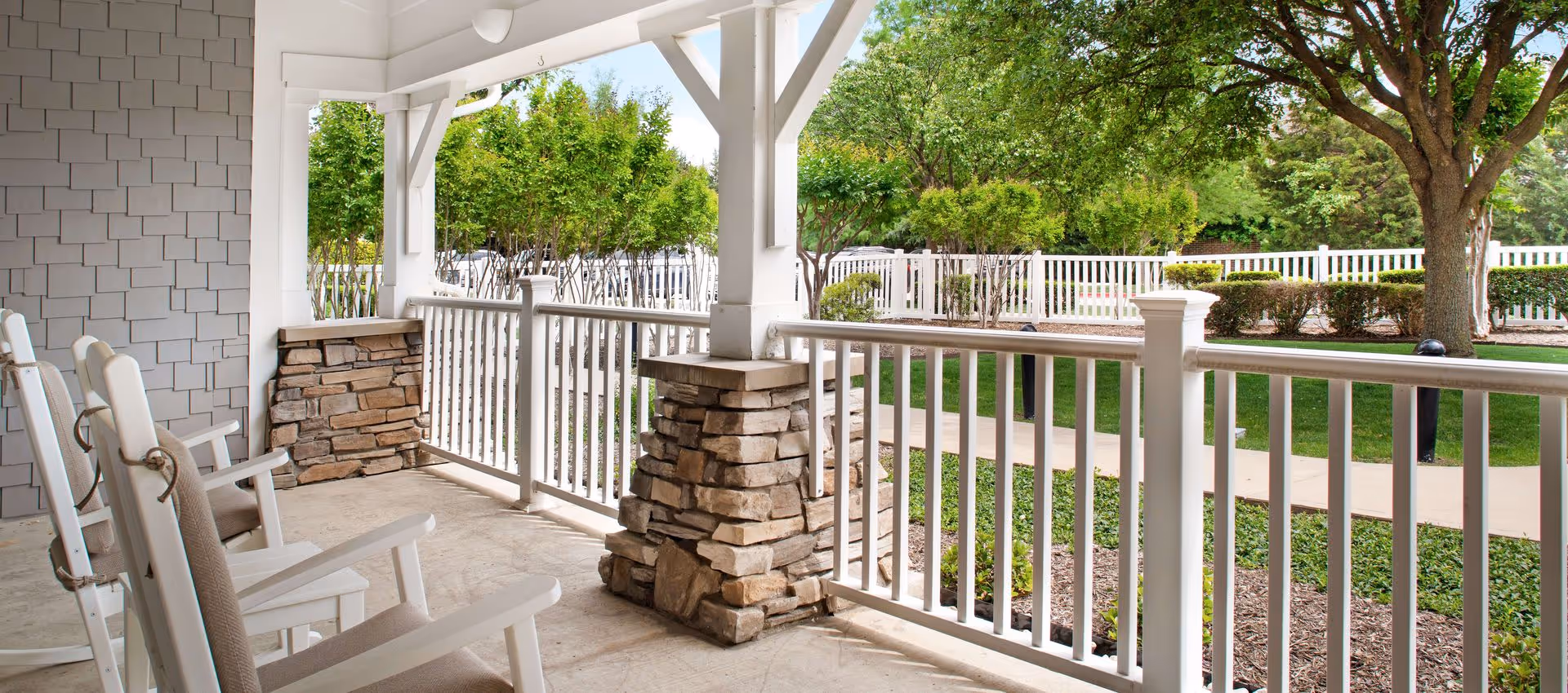 Covered porch area with white rocking chairs and stone pillars, overlooking a landscaped garden with trees, bushes, and a white fence.