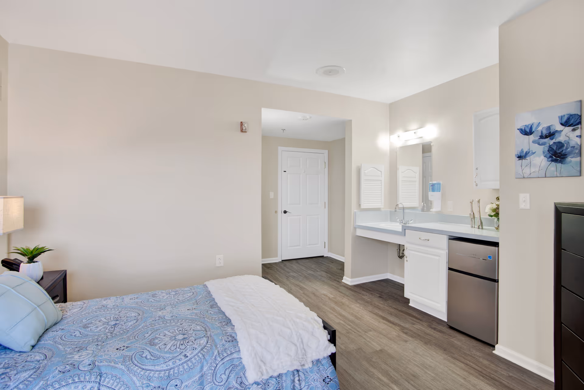 A bright and clean bedroom in a senior living facility with a bed covered in a blue patterned bedspread and a white throw blanket. There is a nightstand with a lamp and a small plant next to the bed. The room has light-colored walls, wood flooring, and a small vanity area with a sink, mirror, and mini refrigerator. A door is visible in the background.