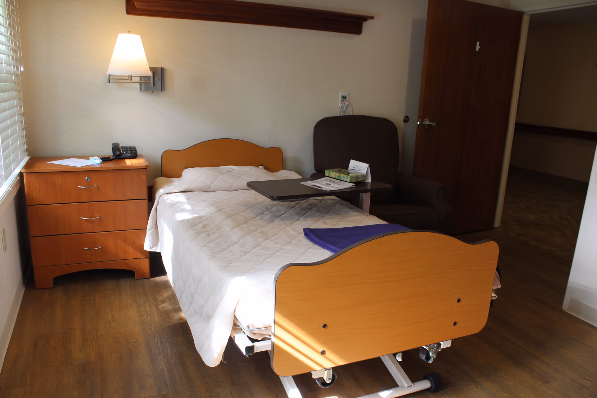 A small, simple bedroom in a senior living facility featuring a single adjustable bed with a light-colored quilt, a wooden nightstand with a telephone and some papers, a wall-mounted lamp, and a dark armchair with a small overbed table holding tissues and a card. The room has wooden flooring and a window with blinds letting in natural light.