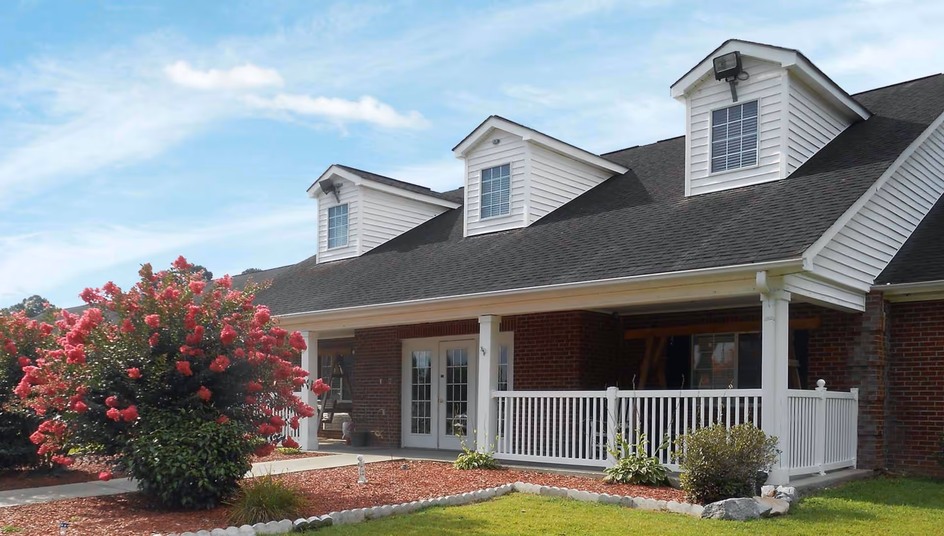 Front exterior of a brick senior living building with a covered porch, white railing, dormer windows, and a large flowering shrub.