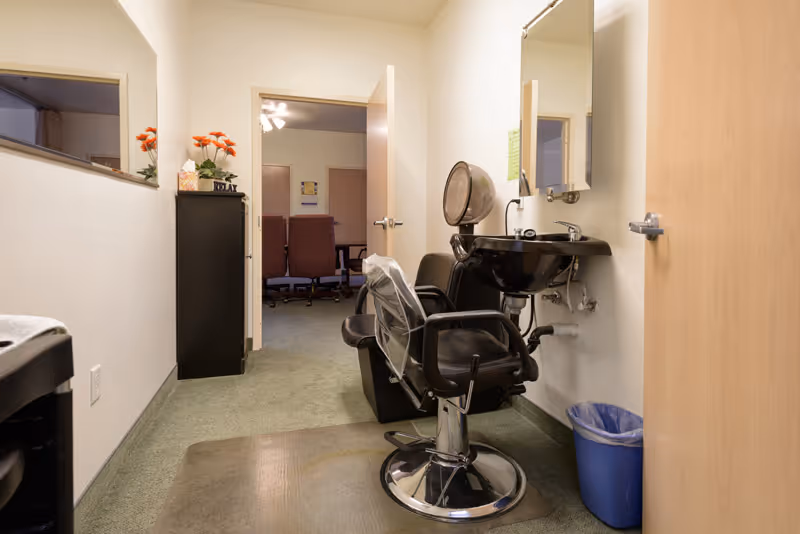 Interior view of a small salon area within a facility, featuring a black salon chair with a plastic cover, a black sink with a mirror above it, a blue trash bin, and a cabinet with orange flowers and a 'RELAX' sign. An open door leads to a room with office chairs and a ceiling fan.