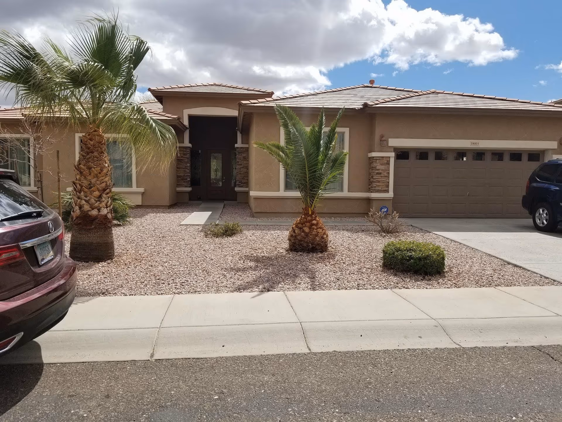 Front exterior view of a single-story house with a tiled roof, two palm trees, and a gravel yard. There are two cars parked on either side of the driveway leading to a double garage door. The sky is partly cloudy.