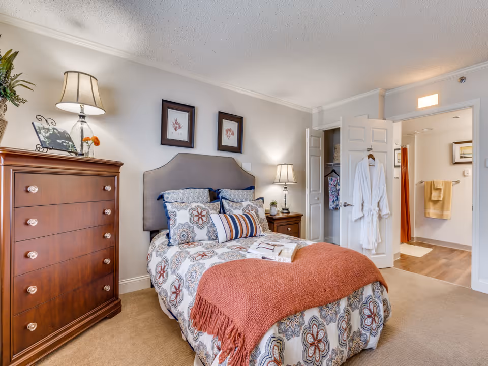 A cozy bedroom with a bed featuring floral patterned bedding and a rust-colored throw blanket. There are two framed pictures on the wall above the headboard. To the left is a wooden dresser with a lamp, a small plant, and a photo frame. To the right, a nightstand with a lamp and a small plant is visible. The door to the bathroom is open, showing a towel hanging on the wall and a shower curtain. A white robe hangs on the bedroom door.