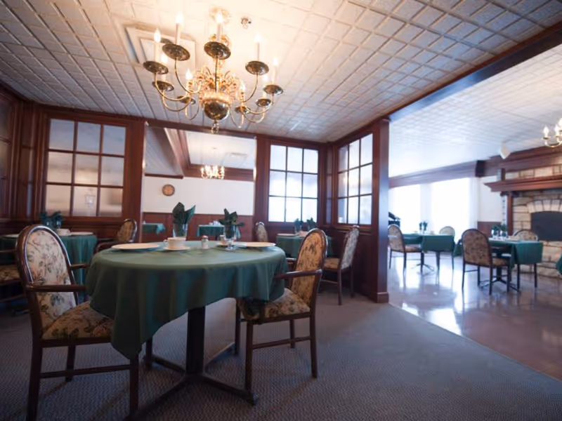 Elegant dining room with round tables draped in green tablecloths, upholstered chairs, chandeliers, and wood-paneled partitions.