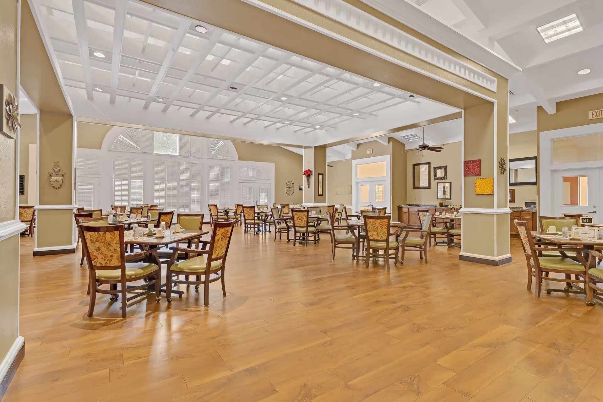 Spacious dining room with multiple wooden tables and chairs arranged neatly on a wooden floor. The room features large windows with white shutters, beige walls, and a white coffered ceiling. Decorative wall art and ceiling fans are visible, creating a warm and inviting atmosphere.