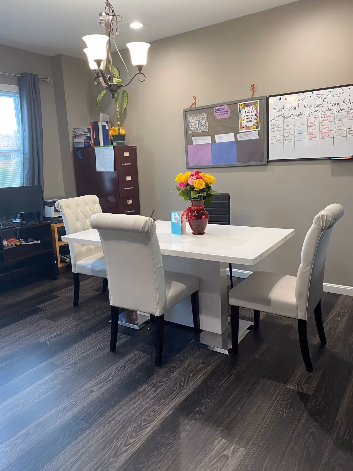 Interior activity/dining room with a white table surrounded by upholstered chairs, a red vase of flowers on the table, and a bulletin/whiteboard on the wall.