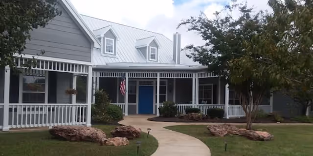 Front exterior view of a single-story building with gray siding, white trim, and a metal roof. The building has a covered porch with white railings and an American flag near the entrance. There is a curved concrete walkway leading to the blue front door, surrounded by landscaping with rocks, bushes, and trees under a partly cloudy sky.
