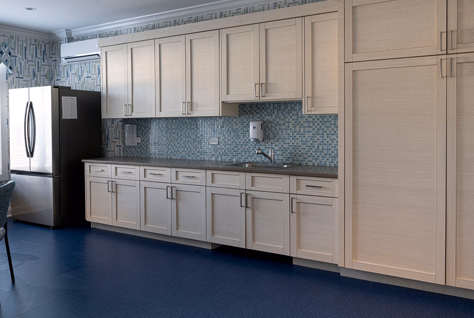 A modern kitchen area with light wood cabinets, a stainless steel refrigerator, a sink with a faucet, and a blue mosaic tile backsplash. The floor is dark blue, and there is patterned wallpaper above the cabinets.