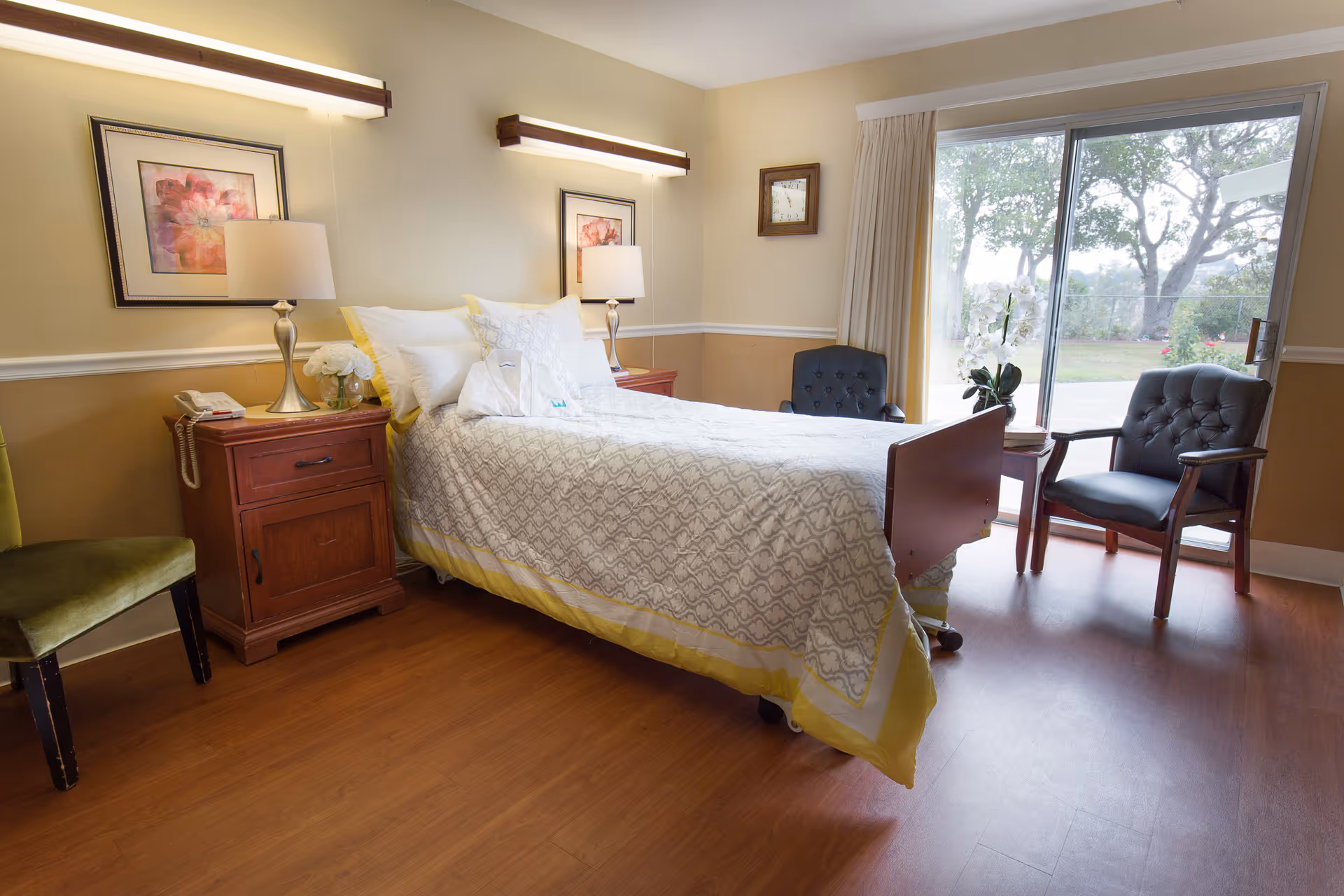 A sunlit resident bedroom with a single bed, wooden nightstand, lamps, chairs, and a sliding glass door to a garden.