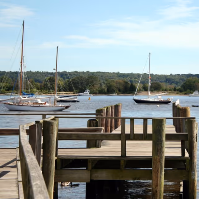 Wooden dock extending over a calm body of water with several sailboats anchored nearby and a tree-lined shore in the background under a partly cloudy sky.