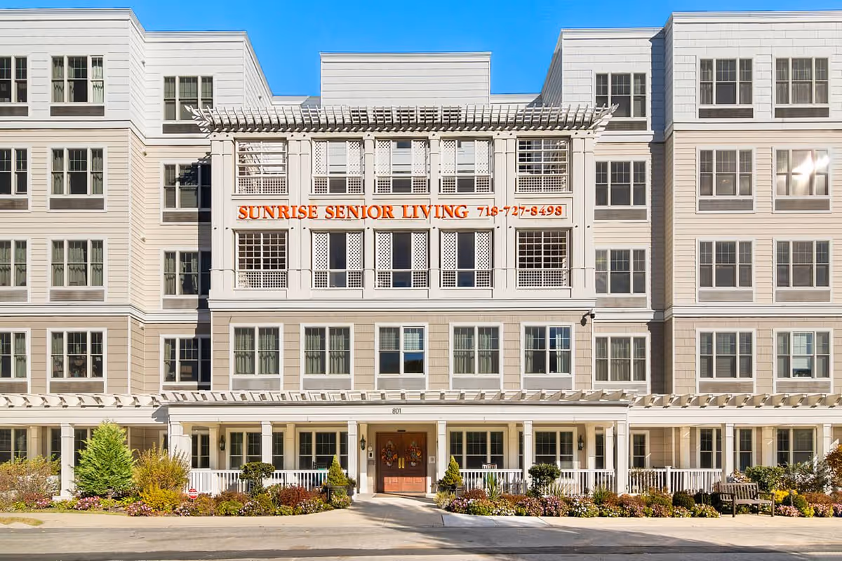 Front exterior of a multi-story Sunrise Senior Living building with a covered entrance, porch and landscaped grounds.