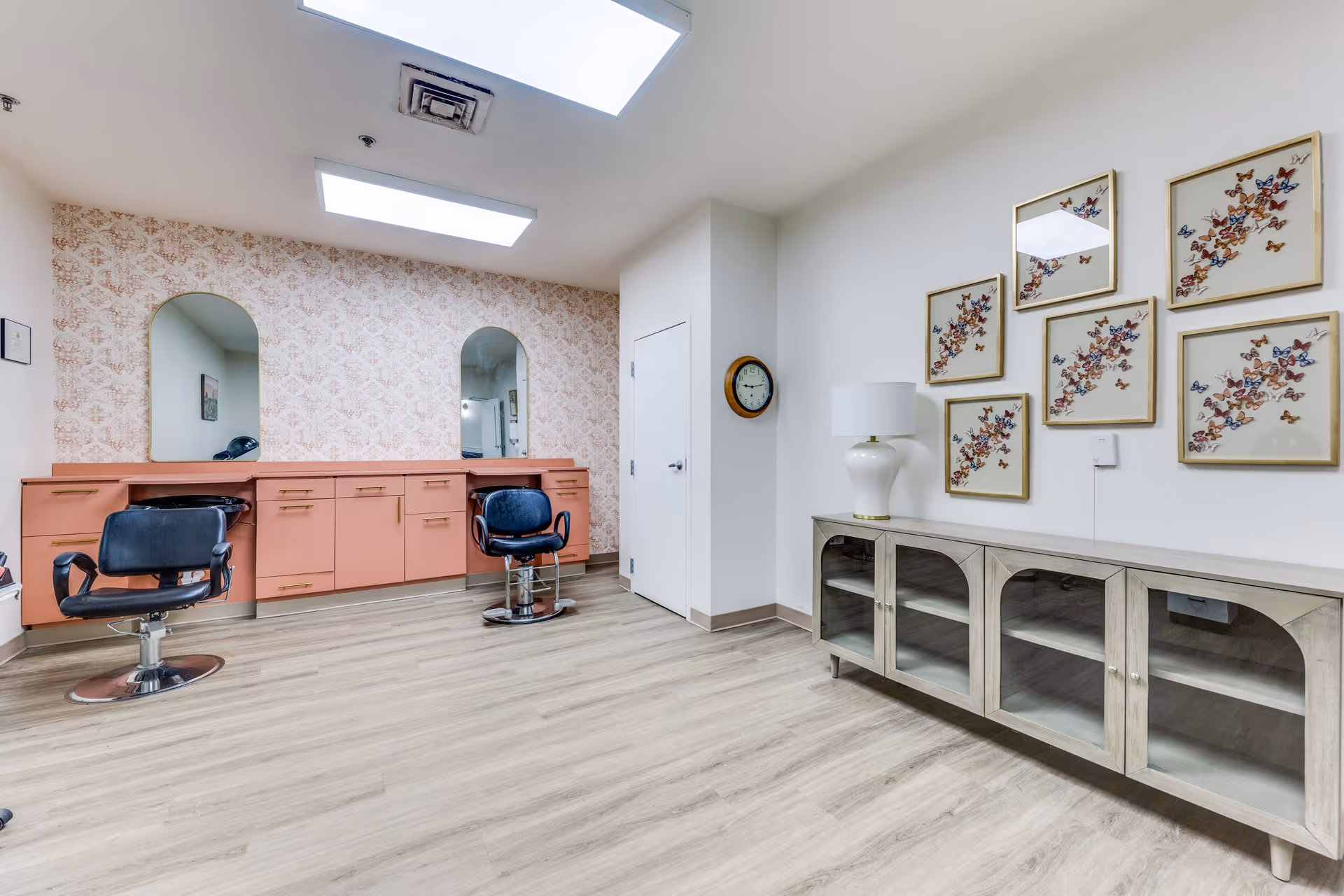 Interior view of a senior living facility room with two black salon chairs in front of a peach-colored counter with cabinets and two arched mirrors. The room has light wood flooring, a white wall with six framed butterfly artworks, a white lamp on a glass-front cabinet, and a wall clock.