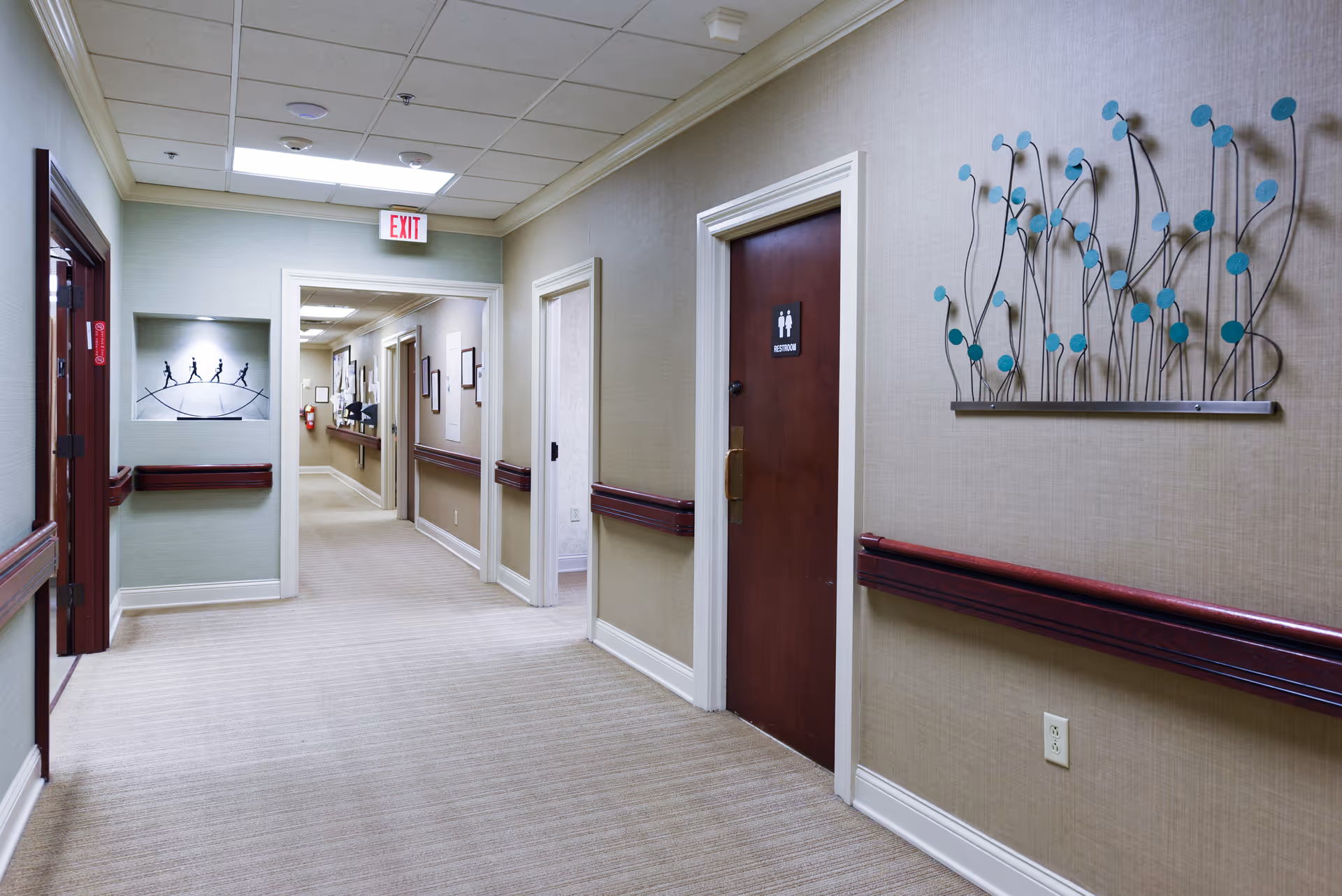 A clean, well-lit hallway in a senior living facility with beige walls and carpeted floor. The hallway features wooden handrails on both sides, a restroom door with a sign, decorative wall art, and an exit sign at the ceiling. The corridor extends into the distance with several doorways and framed pictures on the walls.