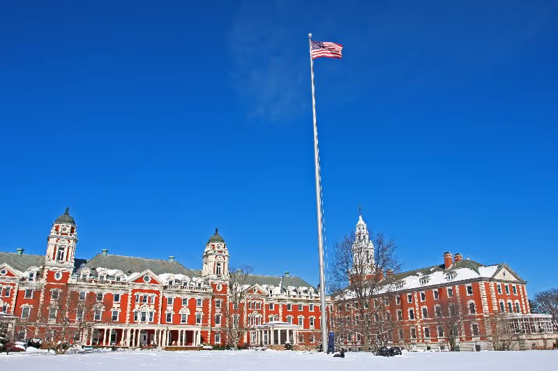 Large historic red brick building with white trim and multiple towers under a clear blue sky, with an American flag flying on a tall flagpole in front and snow covering the ground.