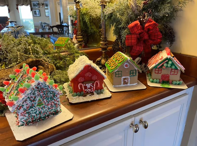 Four decorated gingerbread houses displayed on a wooden countertop with greenery and a red plaid bow decoration in the background. The gingerbread houses are decorated with various colorful candies and icing. A mirror and part of a dining area with chairs and windows are visible in the background.