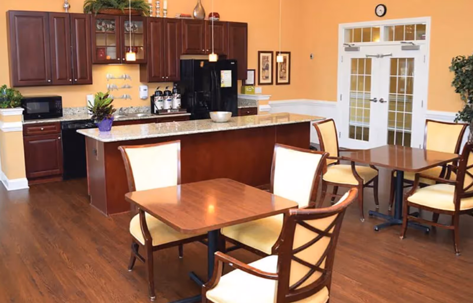 Dining area with wooden tables and upholstered chairs facing a kitchen island with a granite countertop and dark wood cabinets.