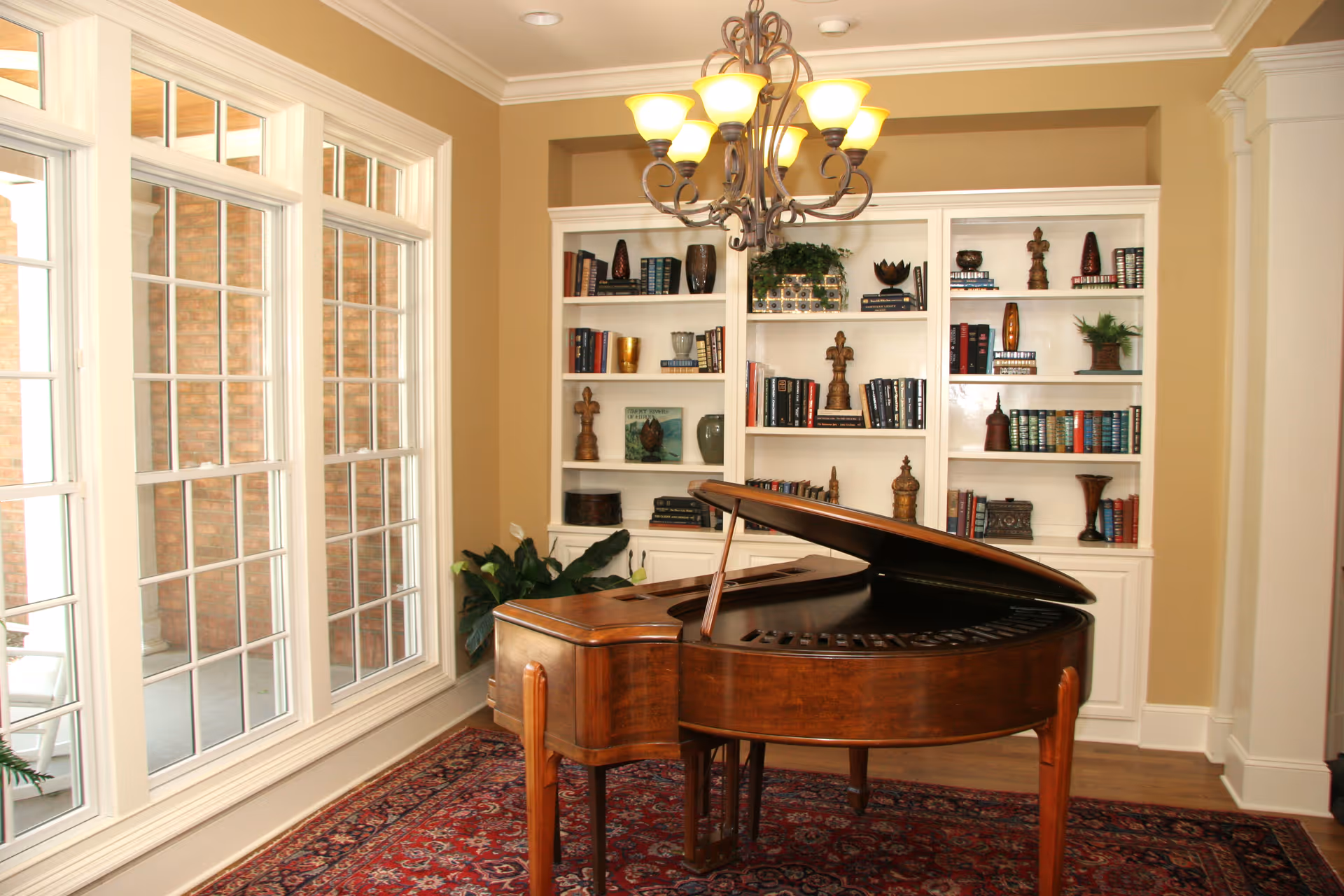 A cozy interior room featuring a wooden grand piano on a patterned red rug, with large windows on the left side letting in natural light. Behind the piano is a white built-in bookshelf filled with books and decorative items. A chandelier with multiple lights hangs from the ceiling.