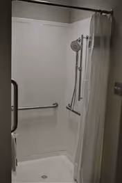 A clean, accessible shower stall with white walls, a handheld showerhead mounted on a vertical sliding bar, a grab bar on the side wall, and a white shower curtain partially drawn to the side.