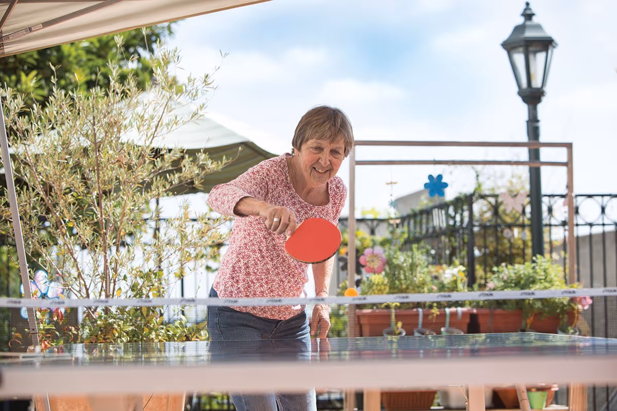 An elderly woman playing table tennis outdoors on a sunny day, surrounded by potted plants and garden decorations, with a street lamp and fence in the background.