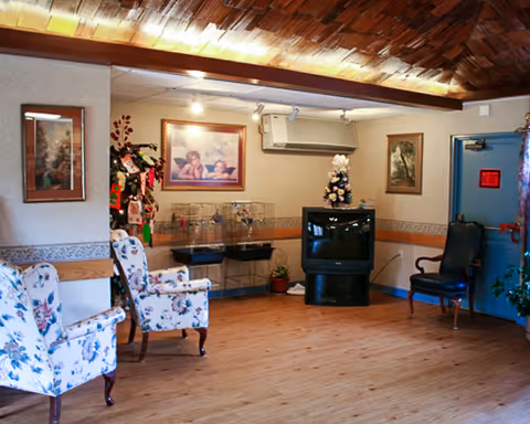 A cozy living room area with wooden flooring and a wooden ceiling. The room features two floral upholstered armchairs on the left, a black TV on a stand in the corner, a small decorated Christmas tree on top of the TV, and a black armchair near a blue door on the right. The walls are adorned with framed pictures and there is an air conditioning unit above the TV. A small plant is placed near the TV stand.