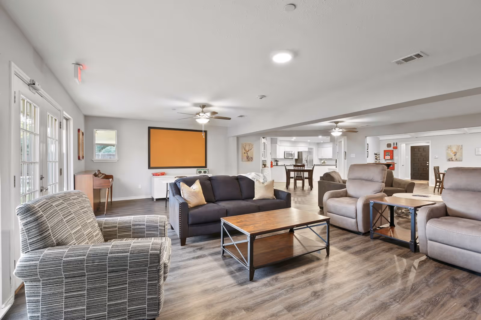 A spacious and well-lit living room area in an assisted living facility featuring a gray patterned armchair, a dark blue sofa with beige pillows, two beige recliners, and wooden coffee and side tables. The room has wood flooring, ceiling fans with lights, and large windows with glass doors letting in natural light. In the background, there is a dining area and a kitchen with white cabinetry and stainless steel appliances.