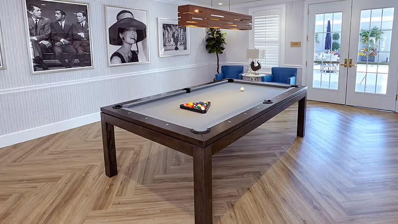 A stylish indoor recreational room with a pool table set up for a game. The room features wood-patterned flooring, three black and white framed photos on the wall, two blue armchairs with a small table and lamp between them, and double glass doors leading outside to a patio area with umbrellas and plants.