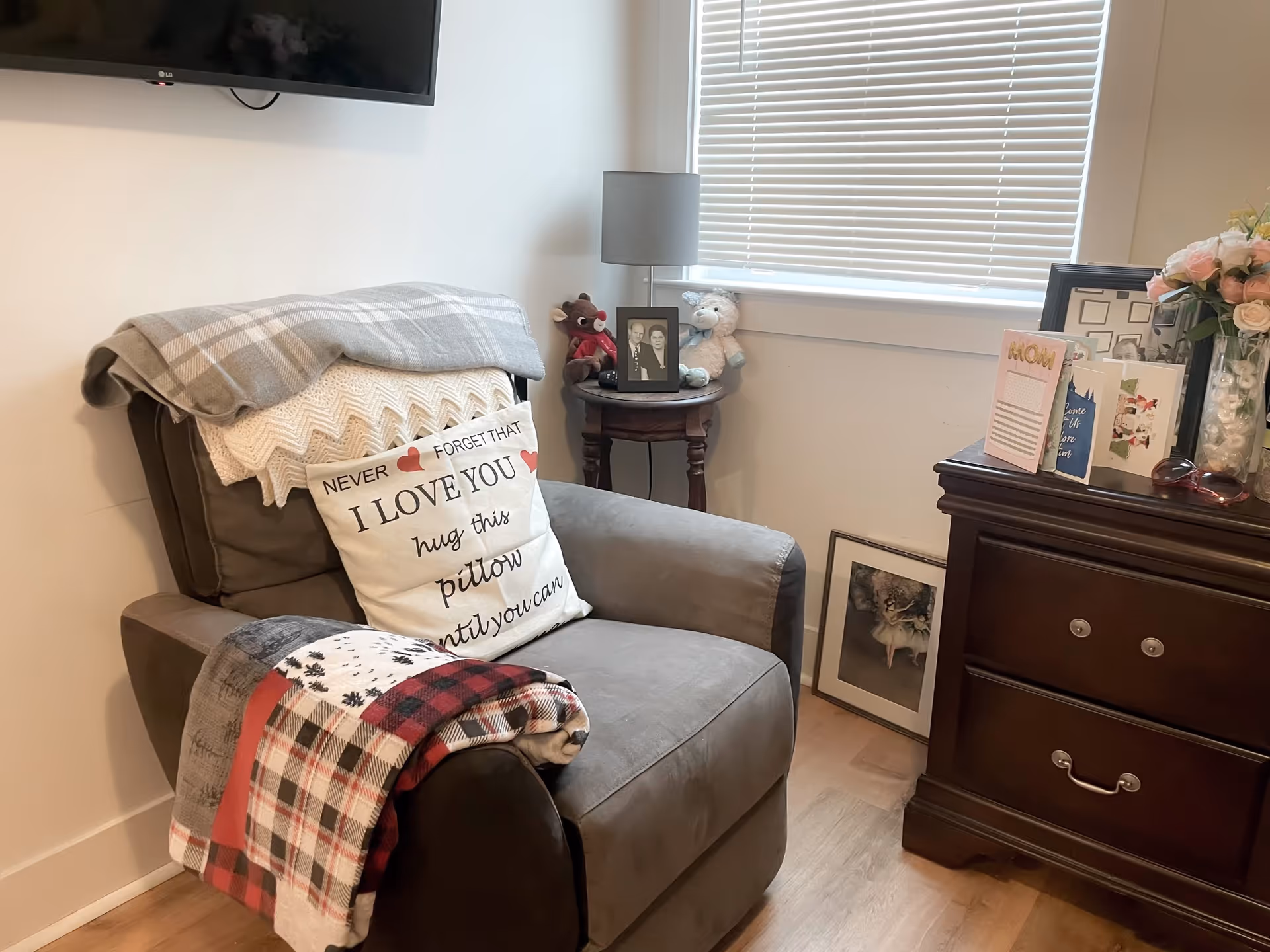 A cozy corner of a room featuring a gray armchair with a decorative pillow that reads 'Never forget that I love you, hug this pillow until you can.' The chair has two folded blankets draped over it. Next to the chair is a small round wooden table with a framed black and white photo, a teddy bear, and a small stuffed animal. A window with closed blinds is behind the table. To the right is a dark wooden dresser with several greeting cards, a vase of flowers, and a framed picture leaning against the wall on the floor.