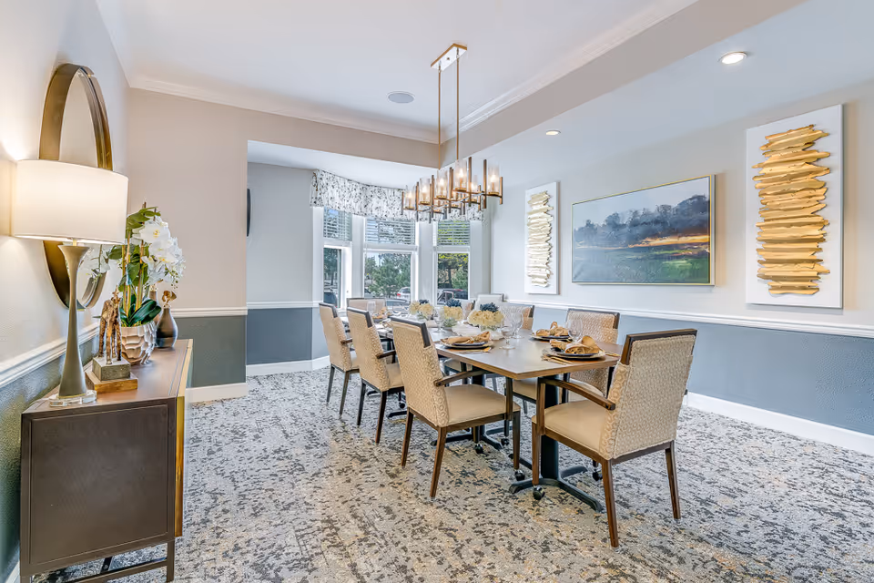 Bright, well-decorated dining room with a long table set for guests, upholstered chairs, a chandelier, wall art, and a sideboard.