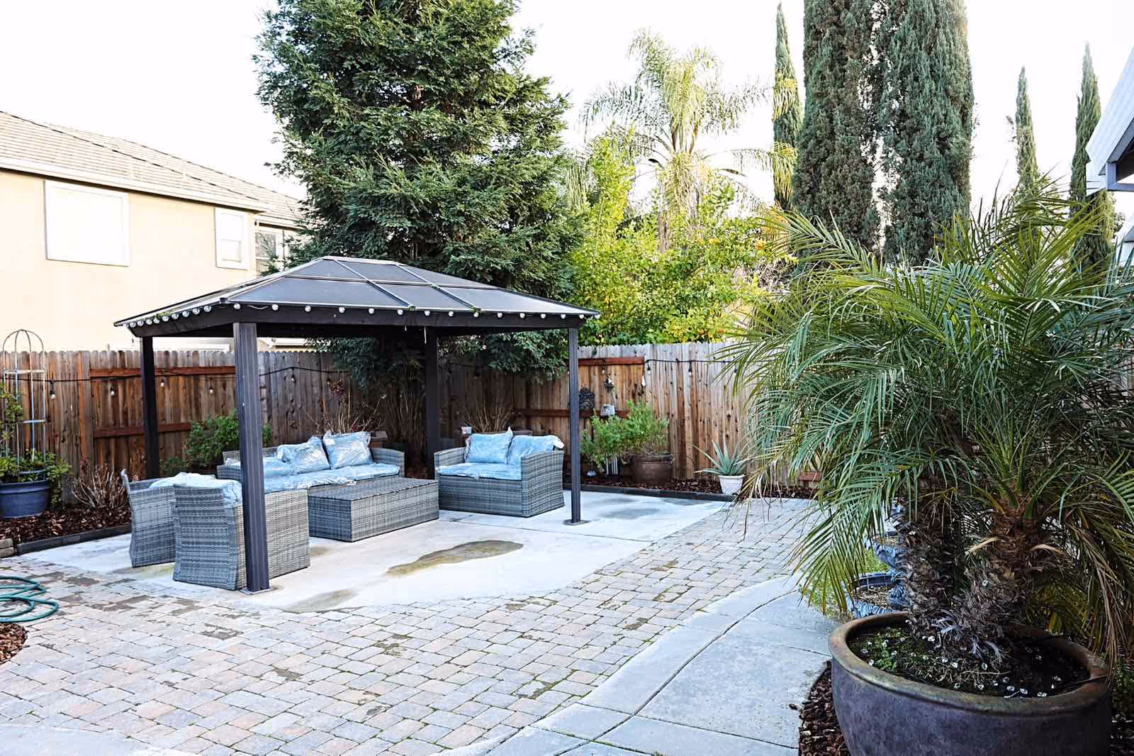 Outdoor patio area with a gazebo covering wicker furniture including a sofa, chairs, and a coffee table. The patio is paved with stone tiles and surrounded by a wooden fence and various green plants and trees.