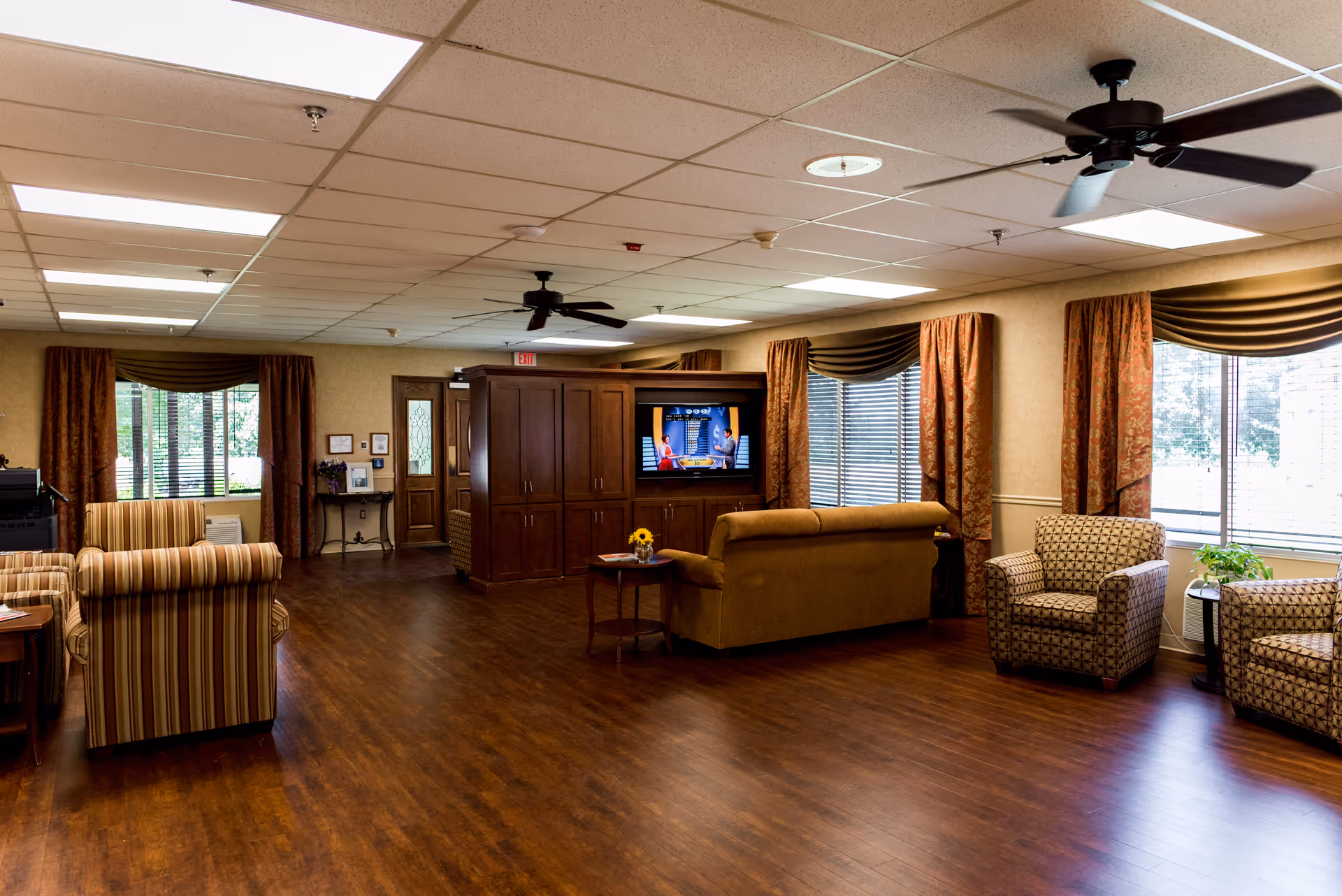 A spacious living room area with wooden flooring, multiple upholstered chairs and sofas arranged around a central wooden entertainment cabinet with a flat-screen TV. Large windows with blinds and curtains allow natural light to fill the room. Ceiling fans and fluorescent lights are mounted on the ceiling.