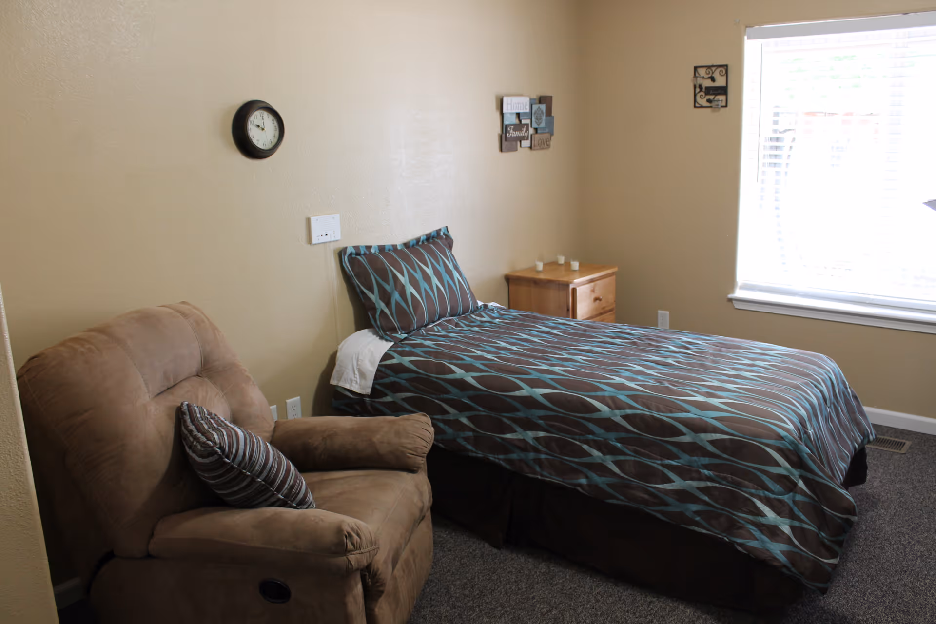 Small furnished bedroom with a single bed covered in brown and teal patterned bedding, a beige recliner, wooden nightstand, wall clock and a window.