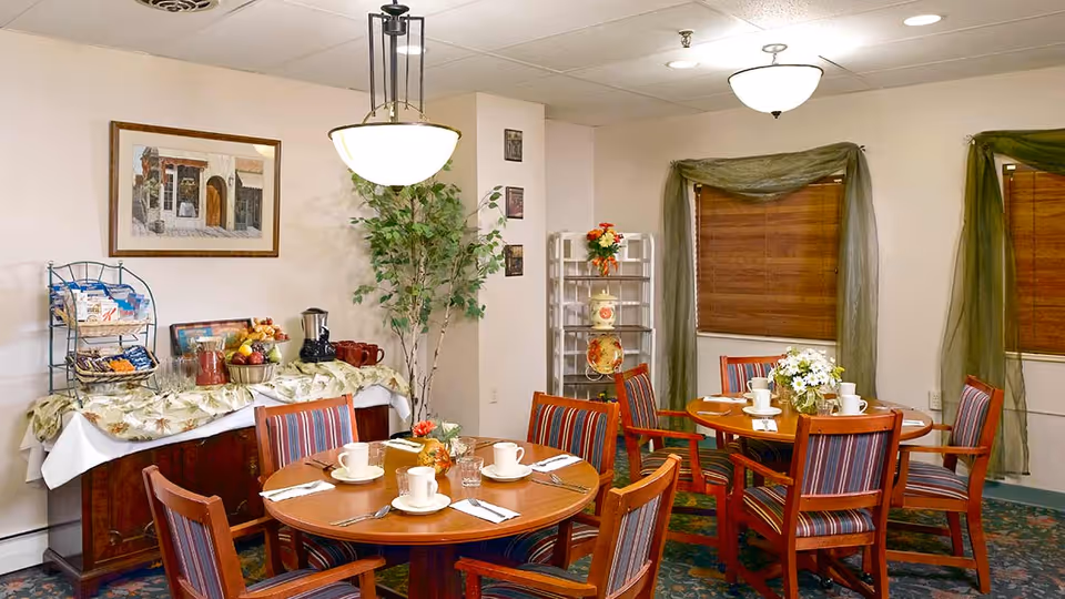 A cozy dining room in a senior living facility with two round wooden tables, each set with white cups, saucers, and utensils. The chairs have wooden frames with striped upholstery. There is a sideboard against the wall covered with a floral tablecloth, holding a variety of snacks, a coffee pot, and a bowl of fruit. The room has beige walls, two windows with wooden blinds and green curtains, a potted plant, and framed artwork on the walls.